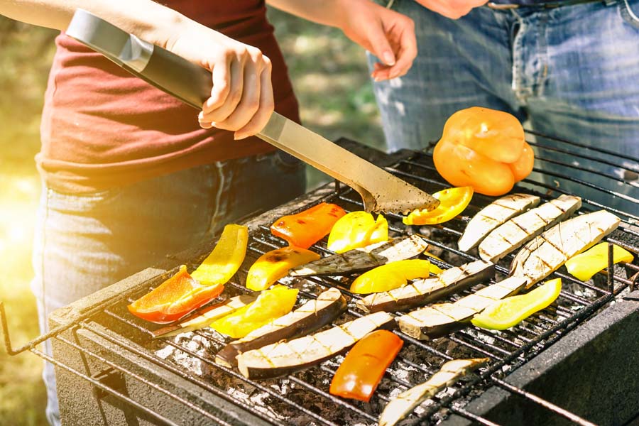 Young woman cooking vegetables for vegetarian barbecue dinner outdoor - Couple grilling peppers and aubergines for bbq - Vegan and healthy lifestyle concept - Soft focus on bottom barbecue tongs