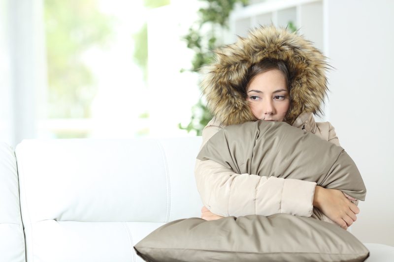 Angry woman warmly clothed in a cold home sitting on a couch; furnace blowing cold air.