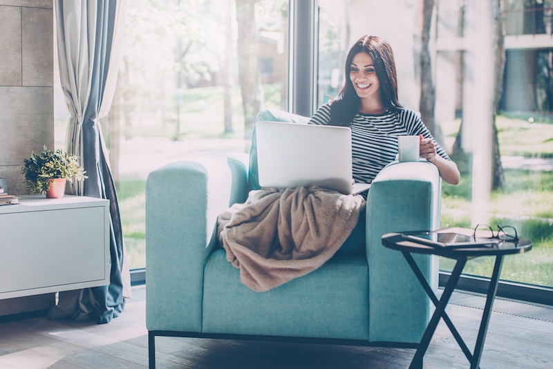 Beautiful young smiling woman working on laptop and drinking coffee while sitting in a big comfortable chair at home